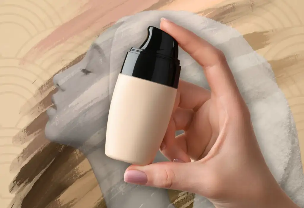 Mano sosteniendo una base de maquillaje líquida sobre fondo artístico beige con rostro femenino desenfocado.
