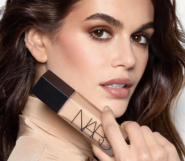 NARS Natural Matte Longwear Foundation maquillaje de larga duración Modelo mostrando el acabado de NARS Natural Matte Longwear Foundation junto al envase del maquillaje de larga duración.