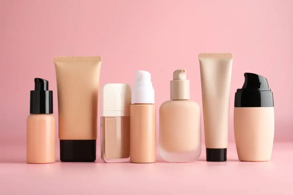 Colección de bases de maquillaje en envases de tubo, frasco y dispensador sobre fondo rosa, representando distintas formulaciones de cosmética facial.