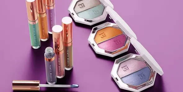 Productos de maquillaje premium con gloss labiales y paletas de sombras multicolor sobre fondo lila.