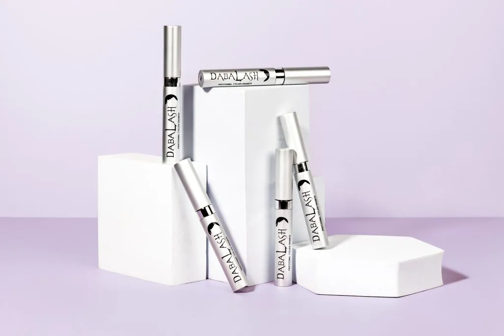 Varios envases del sérum Dabalash Eyelash Enhancer sobre pedestales blancos en una composición de estudio con fondo lila claro.