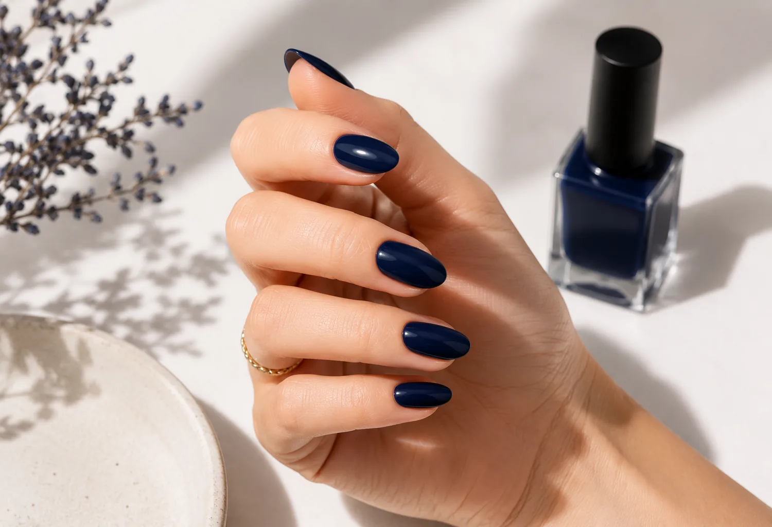 Mano con uñas de color azul marino brillante y acabado glossy junto a un esmalte en un ambiente minimalista iluminado con luz natural.