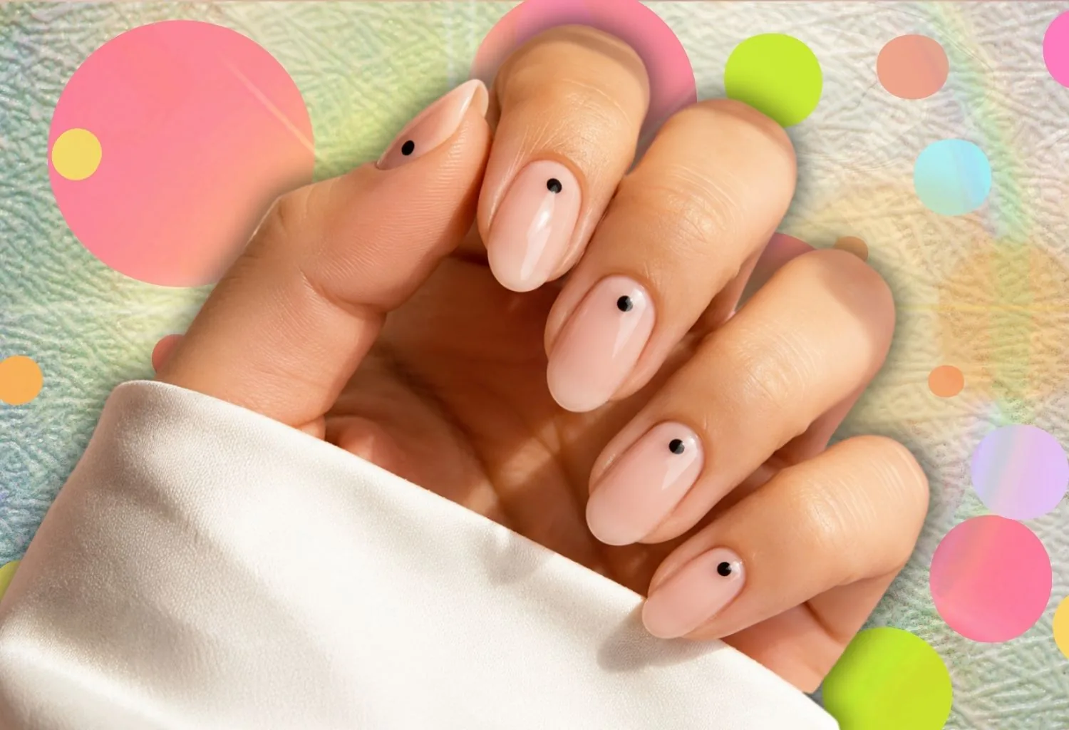 Manicura polka dot minimalista con pequeños lunares negros sobre uñas nude brillantes y fondo colorido de inspiración retro.